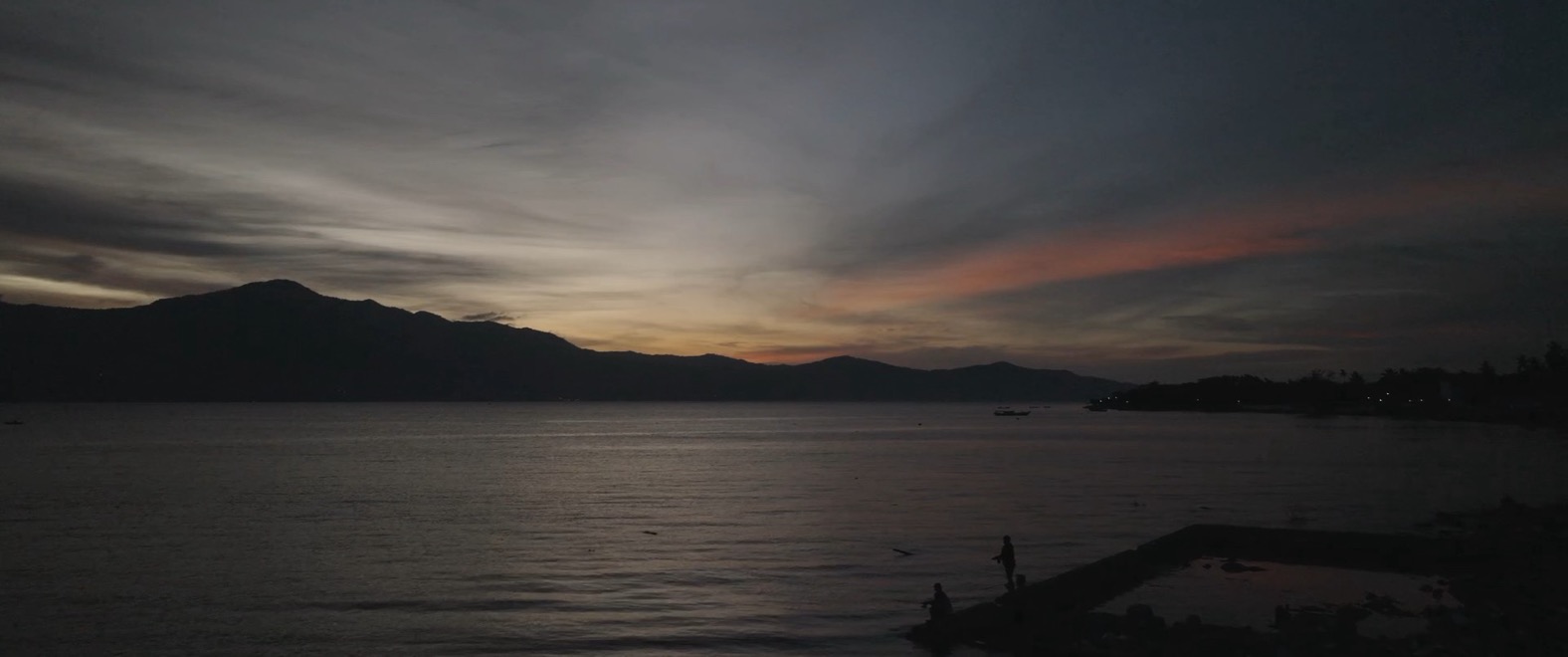 A frame from the reel: two silhouetted figures fishing from a jetty at dusk against layered mountains